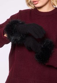 Mujer con guantes de punto negros con puños de pelaje negro sobre un suéter rojo oscuro, ajustándose un guante.
