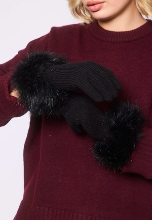 Kvinde iført sorte strikkede handsker med fluffy sorte manchetter over en mørkerød sweater, mens hun justerer den ene handske.