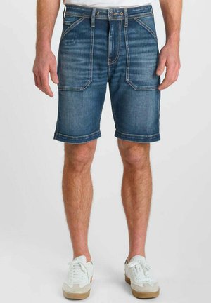 Man draagt halflange blauwe denimshorts met grote voorzakken en witte sneakers tegen een effen achtergrond.