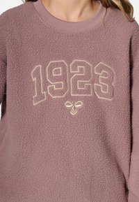 Fleece sweatshirt in mauve with embossed tan numbers "1923" centered on the chest. Soft texture, ribbed crew neckline.