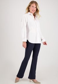 Vrouw in een witte blouse met knopen, marineblauwe gestreepte broek, beige hakken, gouden creolen, glimlachend, staand tegen een effen achtergrond.