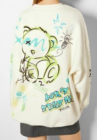 Sudadera grande de color crema con una obra de arte estilo graffiti colorida, que incluye un oso, mariposas y detalles de texto en azul y verde.