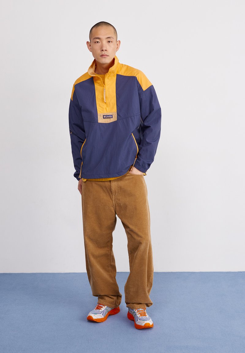 Chaqueta cortavientos azul marino y amarilla con cremallera de un cuarto al frente y bolsillos laterales, combinada con pantalones de pana caqui y zapatillas multicolor.