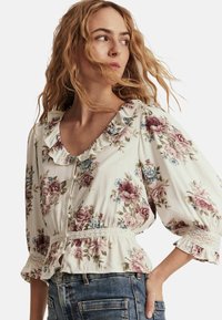Blomstret bluse i lys cremefarvet stof, med rynket halsudskæring, puffede ærmer og en smocket talje med spidskant.
