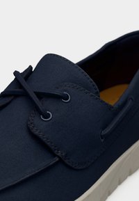 Chaussure bateau en toile marine avec lacets ronds, coutures ton sur ton et empeigne texturée. Dispose d'une semelle contrastée de couleur claire.