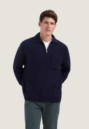 Homme portant un pull zippé bleu marine avec une poche poitrine et un pantalon vert, debout les mains derrière le dos devant un fond clair.