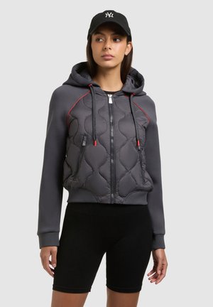 Femme portant une casquette de baseball noire, un sweat à capuche matelassé gris zippé avec des accents rouges, et un short de cycliste noir, sur un fond clair uni.