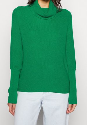 Personne portant un pull vert vif à manches longues avec un col roulé ample et un jean bleu clair sur un fond clair uni.