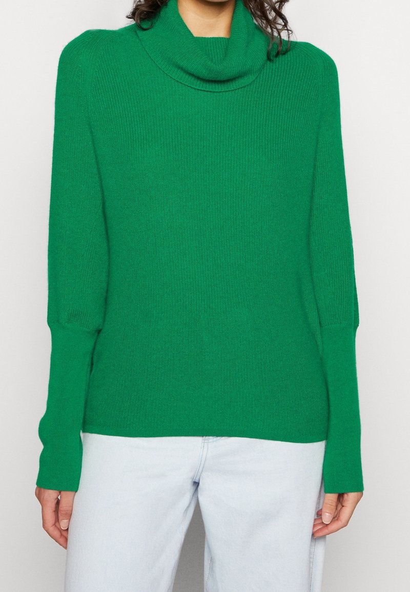 Personne portant un pull vert vif à manches longues avec un col roulé ample et un jean bleu clair sur un fond clair uni.