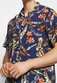 Květinová košile s krátkým rukávem v námořnické modři s živými oranžovými květy a zelenými listy. Dokončená knoflíky a visačkou.