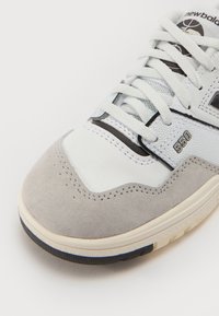 Sapatilhas brancas e cinzas com parte superior em camurça e couro, apresentando detalhes em malha, costuras e uma sola em borracha. Número do modelo 550 visível.