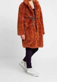 Pelsagtig orange frakke med stor krave, taljebælte og to forlommer, kombineret med stribede navy bukser og hvide høje sneakers.