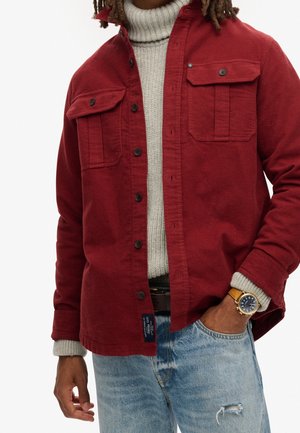 Homme portant une veste rouge boutonnée sur un pull à col roulé gris, un jean bleu déchiré, et une montre-bracelet avec un bracelet en cuir marron.