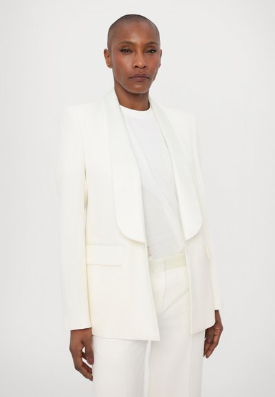 Witte op maat gemaakte blazer met een sjawlrevers, enkele zak en gestructureerde schouders, gecombineerd met een effen wit top en bijpassende broek.