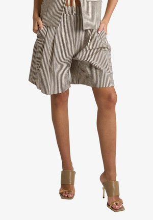 Donna che indossa pantaloncini larghi a righe verticali beige e bianchi con le mani nelle tasche, abbinati a sandali beige con tacco alto e punta aperta.
