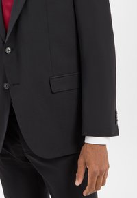 Uomo che indossa un abito nero su misura con giacca abbottonata, polsini della camicia bianca, pantaloni neri e una camicia rossa visibile sotto la giacca.