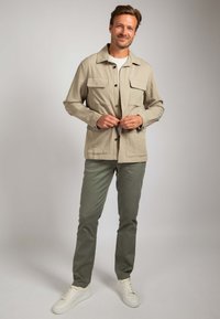 Beige lichtgewicht jas met twee voorzakken, knoopsluiting en een comfortabele pasvorm, gecombineerd met olijfgroene broek en witte sneakers.
