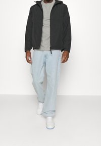 Svart lättviktsjacka med huva, oetad, över en grå t-shirt, i kombination med ljusblå löst sittande jeans och vita sneakers.