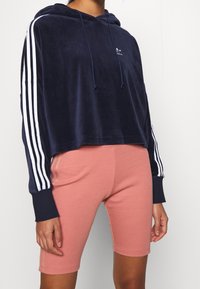 Marinblå velour-hoodie med vita ränder på ärmarna, försedd med huva och dragsko. Matchas med rosa ribbade cykelshorts.