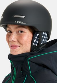 Czarny kask narciarski z logo ROXY, matowe wykończenie, srebrne akcenty. Wyłożony podszewką w kropki, w połączeniu z czarną kurtką z zielonymi wykończeniami.