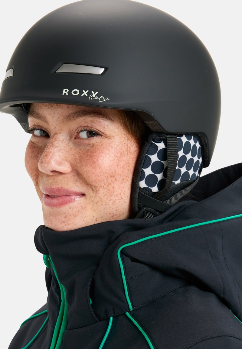 Czarny kask narciarski z logo ROXY, matowe wykończenie, srebrne akcenty. Wyłożony podszewką w kropki, w połączeniu z czarną kurtką z zielonymi wykończeniami.