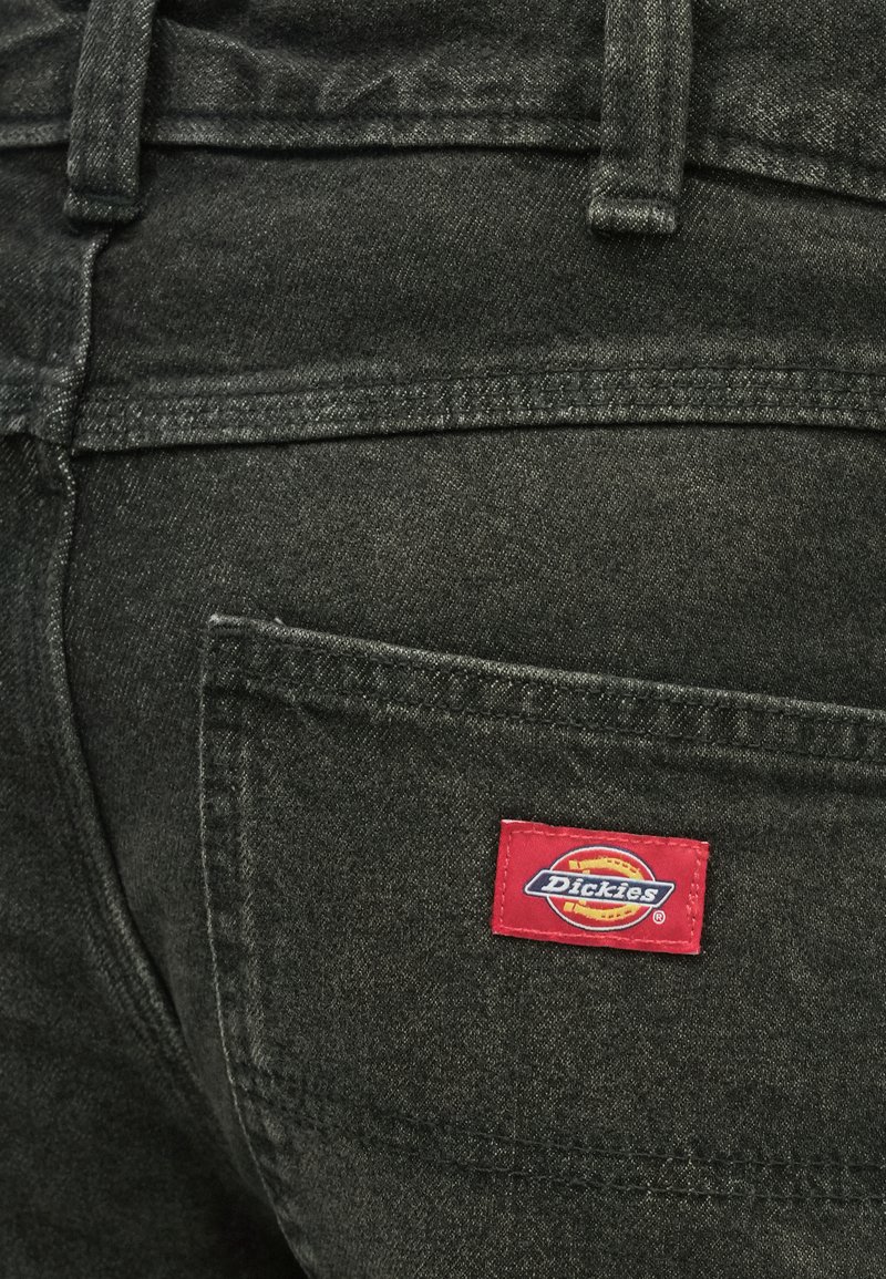 Pantalon en denim noir avec une poche arrière, présentant une étiquette rouge "Dickies". Le matériau a une texture légèrement délavée avec des détails de couture visibles.