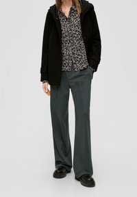 Cappotto nero con cappuccio indossato su una blusa scura a fantasia floreale, abbinato a pantaloni larghi verde scuro e scarpe chunky nere.