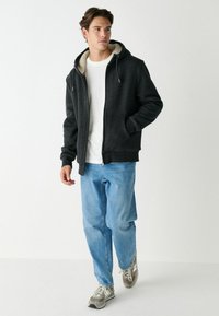 Schwarzer Kapuzenpullover mit einem gefütterten Innenfutter aus Fleece, kombiniert mit hellblauen Jeans und grauen Sneakers. Der Hoodie hat einen Reißverschluss vorne und Taschen.