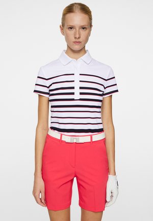 Weißes Polohemd mit Kragen, verziert mit navyblauen und roten Streifen; kombiniert mit leuchtend roten Shorts und einem weißen Gürtel. Eine mit Handschuhen bedeckte Hand ist auf der rechten Seite sichtbar.