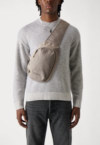 Beige crossover taske med en glat overflade, der har en lynlåslomme og et logo. Båret over en lys grå sweater og mørke jeans.