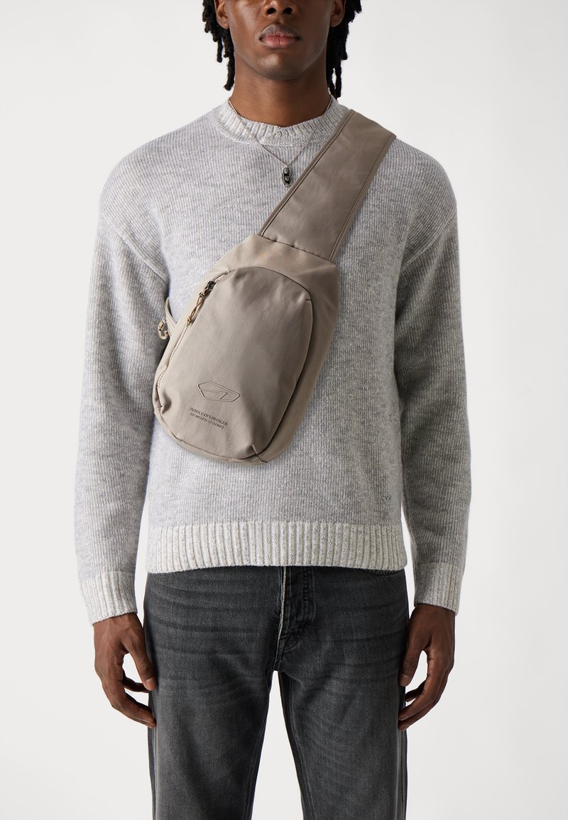 Beige crossbody bag with a smooth texture, featuring a zippered pocket and a logo. Worn over a light grey sweater and dark jeans.