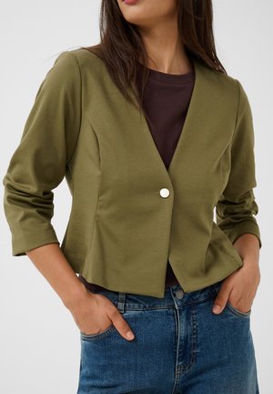 Frau trägt olivgrünen gekürzten Blazer über dunklem Top und blauen Jeans, Hände in den vorderen Taschen, neutraler Hintergrund.
