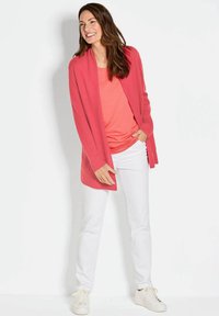 Cardigan corail sur un haut assorti, associé à un pantalon blanc et des baskets blanches. Le cardigan comporte des poignets côtelés et un design ouvert sur le devant.