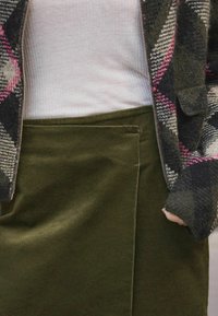 Zöld kordbársony szoknya hajtás részlettel, szürke, rózsaszín és fekete mintázatú zipzáros dzsekivel, alatta világos bordás toppal párosítva.