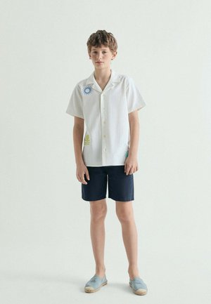 Niño de pie, vistiendo una camisa blanca de manga corta con diseños coloridos de sol, pantalones cortos azul marino y zapatos tipo mocasín azul claro sobre un fondo liso.