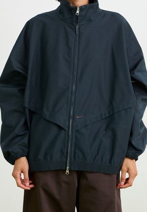 Persoon die een zwart jack met ritssluiting draagt, voorzien van elastische manchetten en een klein Nike-logo, gecombineerd met donkerbruine broek.