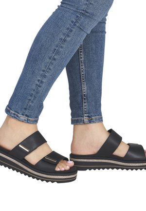 Feet wearing black double-strap platform sandals with gold accents paired with blue fitted jeans, visible against a white background.
