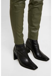 Svarta läderankelstövlar med fyrkantig tå, höga klackar, parat med olivgröna figursydda byxor i konstläder med sömdetaljer.