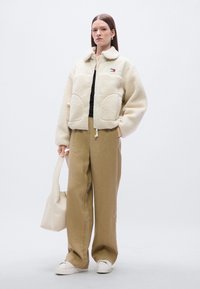 Φλις μπουφάν σε κρεμ χρώμα με δύο υπερμεγέθεις μπροστινές τσέπες και κλείσιμο με φερμουάρ. Συνδυασμένο με μπεζ παντελόνι φαρδιάς γραμμής και λευκά αθλητικά παπούτσια. Κρατώντας μια μπεζ τσάντα.