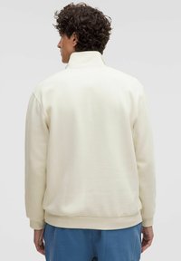 Felpa color crema con colletto alto, polsini a costine e una texture liscia. Progettata con una vestibilità rilassata e senza motivi visibili.