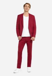 Blazer rosso e pantaloni coordinati in tessuto liscio. T-shirt bianca sotto, completata da sneakers bianche. Dettaglio notevole: accento colorato sul blazer.