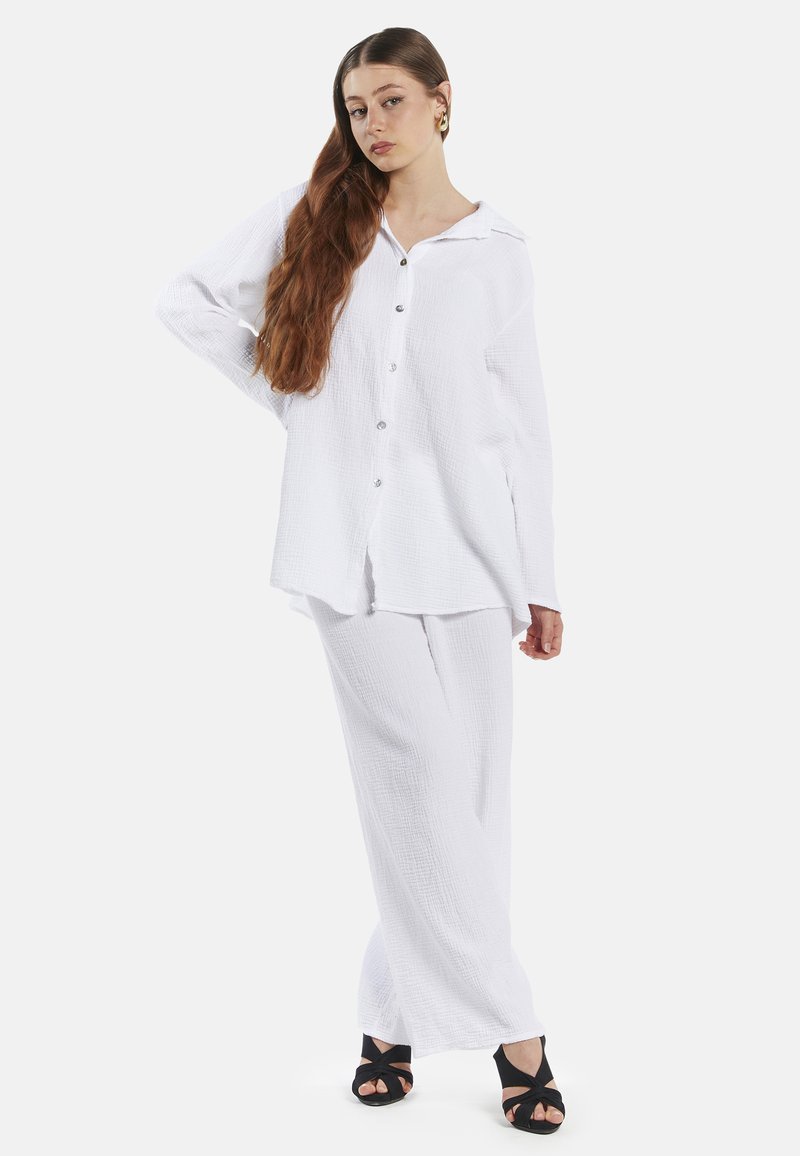 Witte, lichte, textuur shirt met knopen en kraag, gecombineerd met bijpassende losse broek. Model draagt zwarte, sandaaltjes met bandjes.