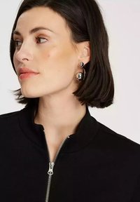 Czarna kurtka zapinana na zamek, z wysokim kołnierzem. Modelka ma na sobie okrągłe srebrne kolczyki o błyszczącym wykończeniu. Krótkie ciemne włosy otaczają twarz.