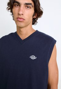 Suéter sin mangas de color azul marino con diseño de pico, textura acanalada y un pequeño logotipo blanco en el pecho.