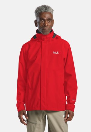 Man draagt een felrode waterdichte jas met capuchon en beige broek, staand tegen een eenvoudige lichte achtergrond.
