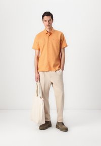 Camisa de botão de manga curta laranja com um bolso no peito, combinada com calças largas bege e calçado castanho, segurando uma bolsa de lona.