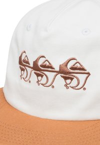Vit keps med en broderad brun design som föreställer berg, kompletterad med en brun skärm. Texturerad tyg med synliga sömnads detaljer.