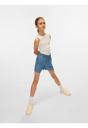 White sleeveless top, blue fabric shorts with elastic waist, cream slip-on shoes, and striped light-colored socks; standing pose.