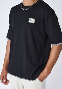 T-shirt nero a maniche corte con righe, caratterizzato da dettagli con cuciture laterali, scollatura rotonda e una patch con logo bianco sul davanti.