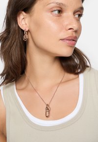 Guldtonet metal smykker med sammenflettede hoops øreringe og en vedhængs halskæde. Halskæden har et unikt knude- og sikkerhedsnålsdesign.